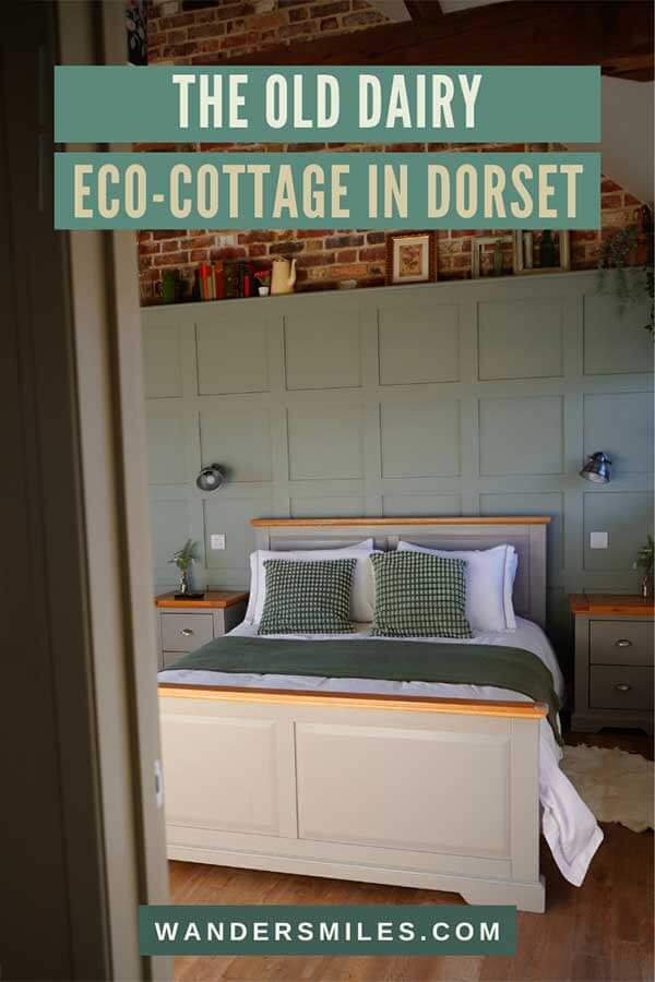 Image of double bed through a door. Sage green wood panelling behind the bed with red brick at the top. Text overlaid text “The Old Dairy Eco Holiday Cottage in Dorset”
