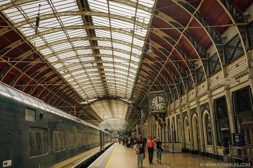 Architecture in Paddington Station, London. The photo shows a train on the left, and arched roof with glass panel, people walking on the platform on the right.