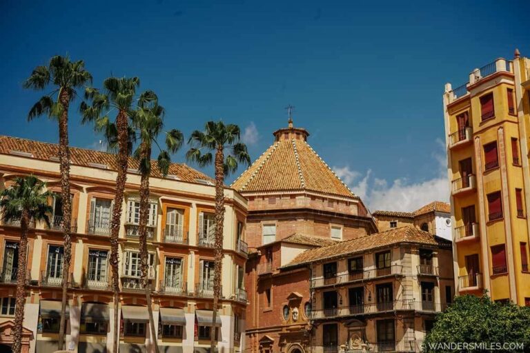 Plaza de la Constitución – in Malaga. Blue sky, palm trees and buildings with a mix of architectural styles, primarily Neoclassical, Baroque, and Andalusian. Feature photo for blog on "Things to do in Malaga"