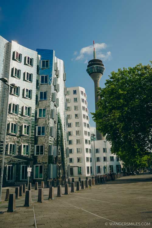 Photo shows two of the Gehry Buildings. One white and one silver organically shaped buildings with the Rhine Tower behind.