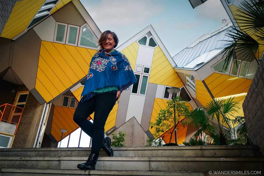 Photo of a white woman (Vanessa from She Wanders Miles) in a blue poncho in front of yellow painted cube houses in Rotterdam