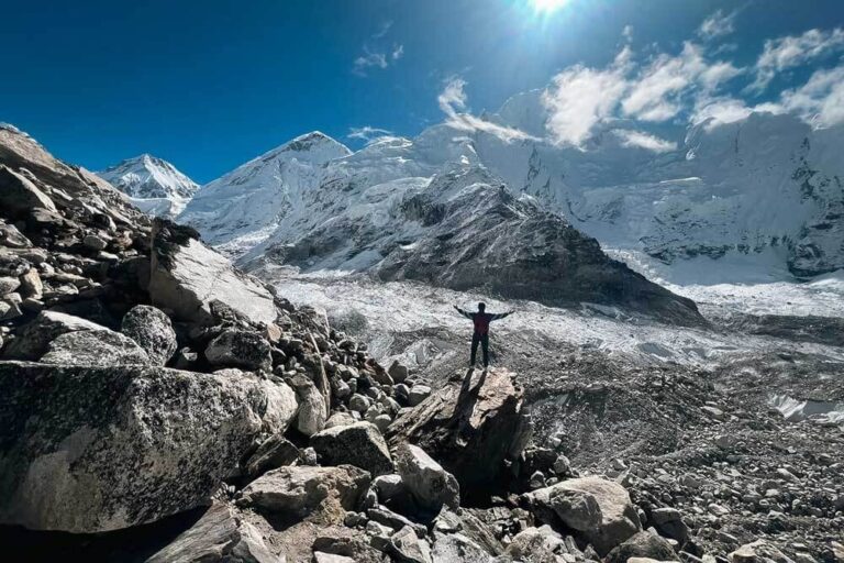 A man standing on a rock high in the Himalayas facing the sun with his arms stretched out. Blue skies and snowy peaks.