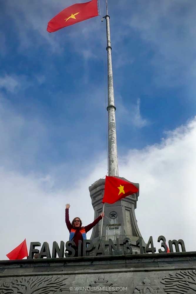 Woman standing with arms raised beside Vietnamese flags at the 3,143 m summit marker on Fansipan mountain under a cloudy sky.