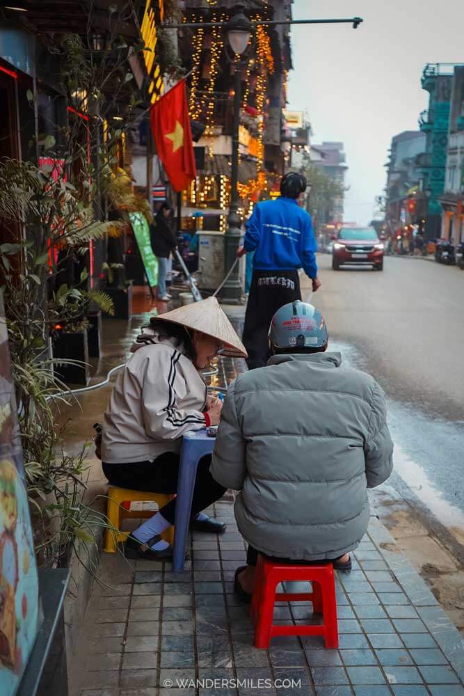Two people sitting on small plastic stools eating at a street food stall on a rainy street in Sapa, Vietnam, with Vietnamese flags and warm lights hanging from nearby buildings