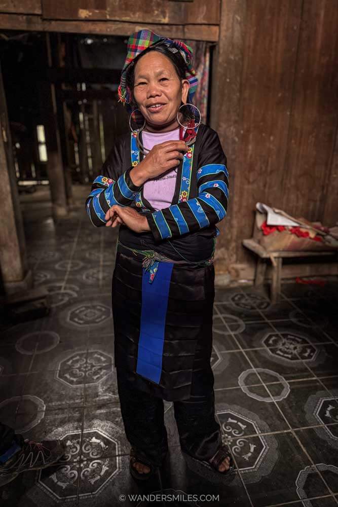 Photo of a txiv neeb, a H&rsquo;Mong shaman, standing dressed in black with blue brocade trims to jacket, and bright plaid head scarf