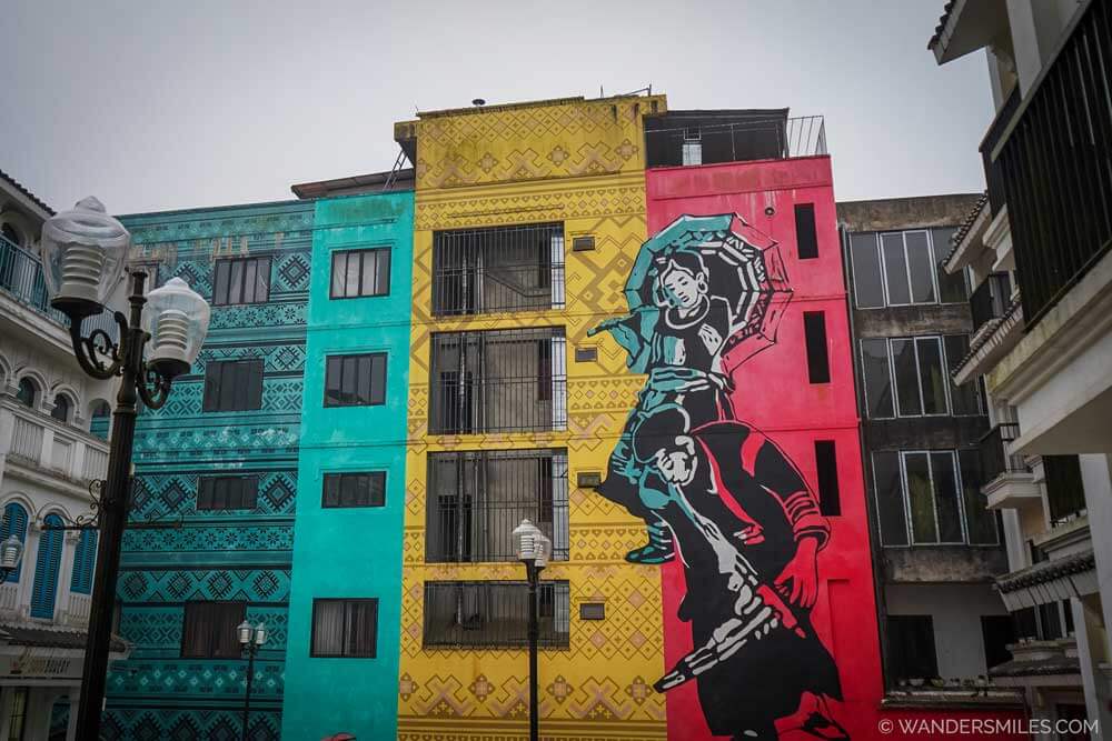 A colourful mural of an ethnic minority woman with a child painted across bright turquoise, yellow, and red buildings in Sapa, Vietnam.