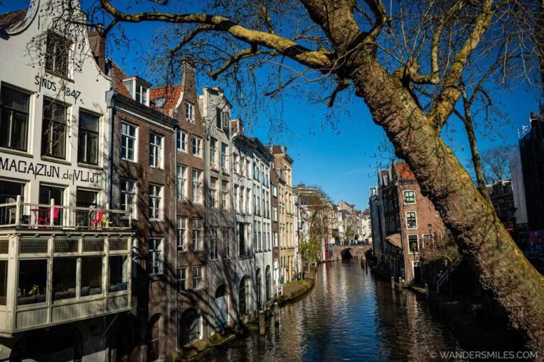 Old warehouses by the canal along Oudegracht in Utrecht – with blue skies and a tree reaching over in the foreground. Feature photo for blog about a "long weekend in Utrecht"
