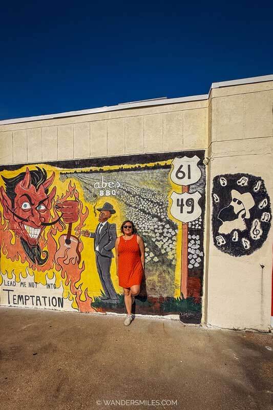 A woman in orange dress standing against the Robert Johnson Temptation mural Abe's BBQ, Crossroads in clarksdale, where he is selling his soul to the devil
