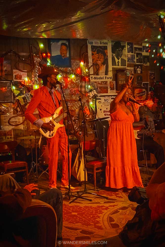 Live Music in Red’s Juke Joint, Clarksdale, Mississippi. A woman in a red dress signing and a man in a red suit playing guitar surrounded by people sitting and watching