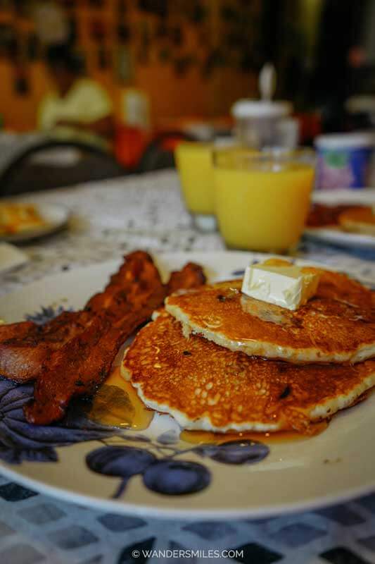 Breakfast at Our Grandma's House of Pancakes. The photo shows blueberry pancakes, butter, maple syrup, and syrup, and crispy bacon on the side. Orange juice in the background.