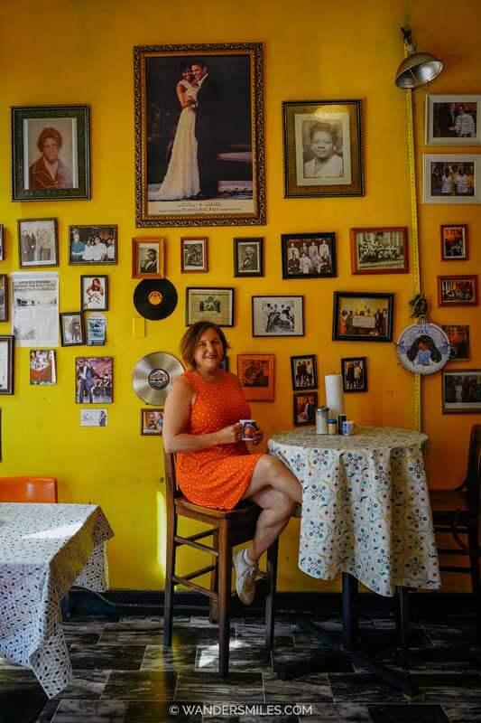 A woman in an orange dress is sitting drinking coffee at Our Grandma's House of Pancakes, Clarksdale. Yellow background with family photos on the wall.