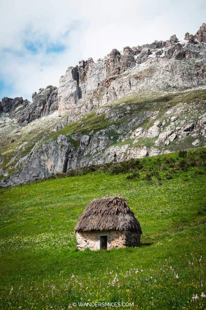 A traditional teito thatched-roof hut with green pastures and high cliffs, in Valle de Lago, Asturias