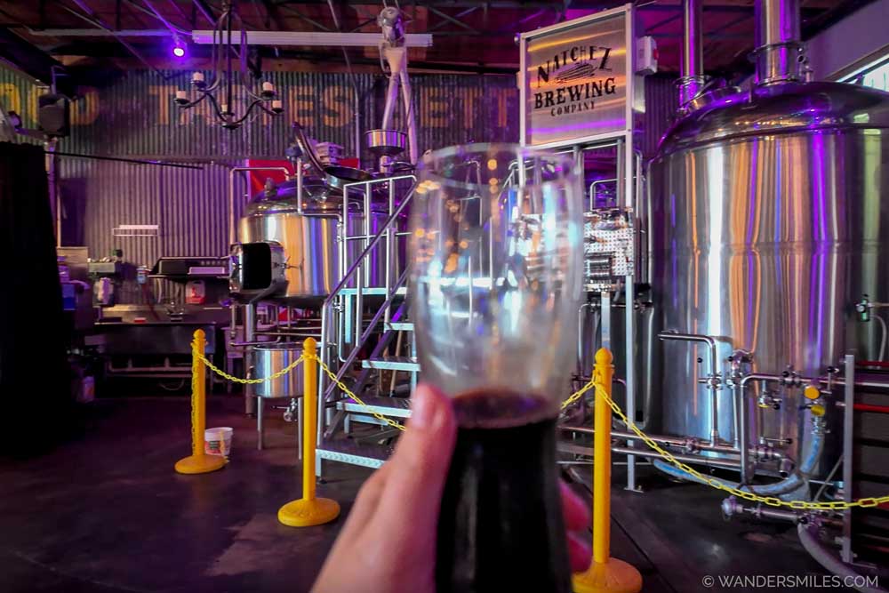 Natchez Brewing Co – A beer held up in front of the silver brewing vats in a warehouse style venue